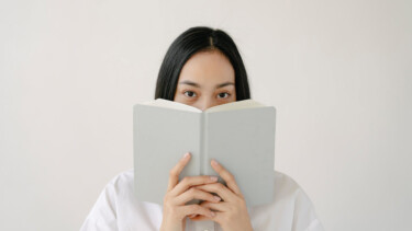 Frau hält sich Buch vor Gesicht [Quelle: pexels.com, John Diez]