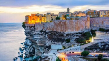 Die Stadt Bonifacio auf Korsika im Sonnenuntergang. Man sieht die halb beleuchtete Stadt an einer Felsklippe, darunter das Meer.