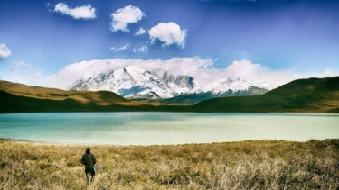 Ein Wanderer steht vor einem See. Im Hintergrund sieht man die Bergkette im Torres Del Paine Nationalpark unter einigen Wolken verborgen.