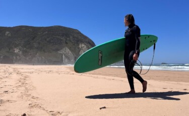 Nike Rendenbach trägt ein Surfboard den Strand entlang.