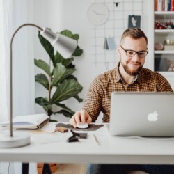 Junger lächelnder Mann mit Brille sitzt am Schreibtisch und schaut auf den Laptop [Quelle: pexels.com]