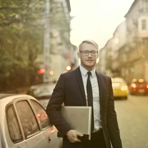 Entschlossener lächelnder Geschäftsmann mit Laptop auf Straße [Quelle: pexels.com]