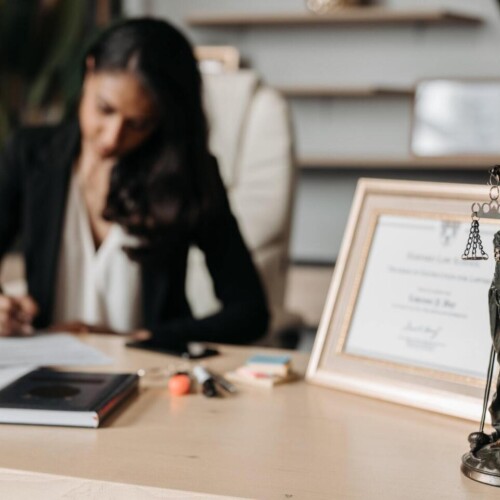 Eine Anwältin sitzt an einem Schreibtisch und unterschreibt ein Dokument. Auf dem Schreibtisch steht ein eingerahmtes Zertifikat neben einer Justizia-Statue. [Quelle: pexels.com]
