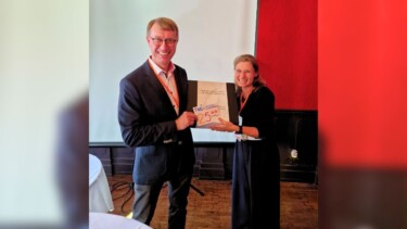 Ein Mann im Anzug und eine Frau im Klein halten ein Geschenk hoch und lächeln in die Kamera