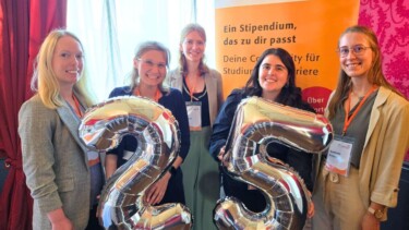 Fünf Frauen stehen in einer Gruppe zusammen und halten zwei Luftballons hoch, die die Zahl 25 bilden.