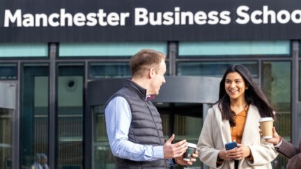 Drei Studierende stehen mit Kaffee-Bechern in der Hand vor dem Eingang der Alliance Manchester Business School und unterhalten sich.