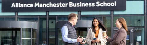 Drei Studierende stehen mit Kaffee-Bechern in der Hand vor dem Eingang der Alliance Manchester Business School und unterhalten sich.