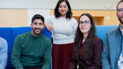 Fünf Studierende sitzen auf einer blauen Couch und lachen in die Kamera
