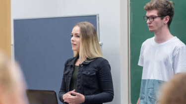 Studierende, eine Frau und ein Mann, präsentieren im Seminarraum einen Vortrag
