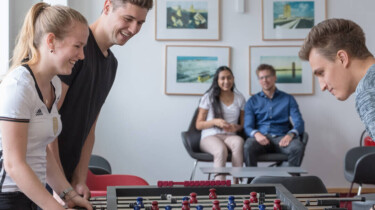 Drei Studierende, zwei Männer eine Frau, spielen an einem Tischkicker gegeneinander. Im Hintergrund sitzen zwei Studierende und betrachten das Spiel.