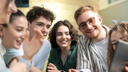 Sechs Studierende, vier Frauen, zwei Männer stehen in einer Gruppe zusammen und schauen gemeinsam auf einen Laptop.