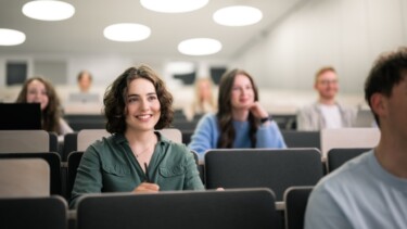 Eine Studentin sitzt in Hörsaal und lächelt.