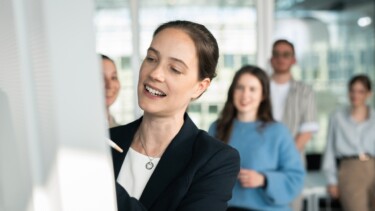 Eine Dozentin steht an einer Flipchart und erklärt den Studierenden im Hintergrund einen Sachverhalt.