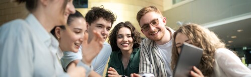 Sechs Studierende, vier Frauen, zwei Männer stehen in einer Gruppe zusammen und schauen gemeinsam auf einen Laptop.