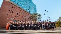 Studierende werfen Absolventenhüte in die Luft und stehen unter der Elbphilharmonie.