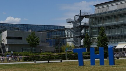 Campus der TH-Ingolstadt. Im Hintergrund das Hochschulgebäude, im Vordergrund die drei Buchstaben "T", "H" und "I" in blau auf einer Wiese stehend.