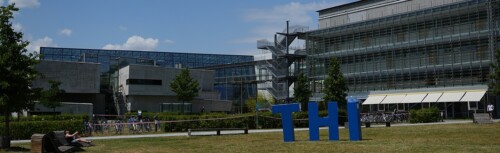 Campus der TH-Ingolstadt. Im Hintergrund das Hochschulgebäude, im Vordergrund die drei Buchstaben "T", "H" und "I" in blau auf einer Wiese stehend.