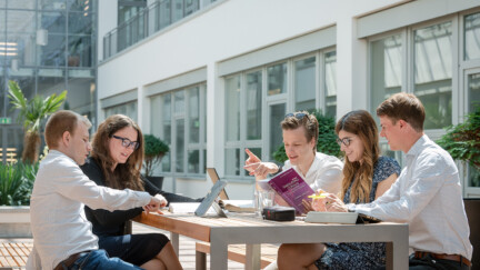 Studierende sitzen im Freien an einem Tisch und lernen gemeinsam. Auf dem Tisch stehen Laptops und liegen Bücher.