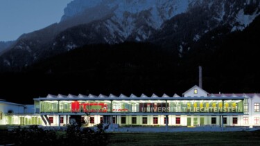 Das Hauptgebäude der Universität Liechtenstein bei Nacht. Im Hintergrund schneebedeckte Berge.