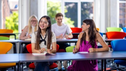 Gruppe von Studierenden in einem modernen Seminarraum mit bunten Stühlen, die lächelnd miteinander sprechen und Notizen machen.