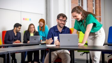 Gruppe von Studierenden arbeitet gemeinsam an Laptops in einem Seminarraum, zwei Personen lachen und schauen auf einen Bildschirm.