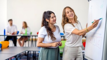 Zwei Studentinnen präsentieren an einem Whiteboard mit Diagramm, während andere im Hintergrund diskutieren.