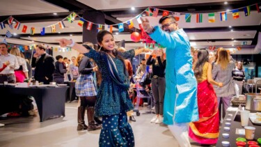 Studierende in traditionellen Outfits tanzen fröhlich bei einer internationalen Feier mit Länderflaggen im Hintergrund.