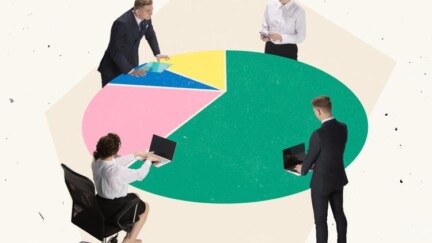 Geschäftsleute stehen und sitzen um einen runden Tisch, der aussieht wie ein Kreisdiagramm. Sie scheinen sich in einem Meeting zu befinden und tippen teilweise auf ihren Laptops.
