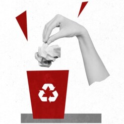 Hand wirft ein zerknülltes Stück Papier in einen Mülleimer, auf dem ein Recycling-Symbol angebracht ist.
