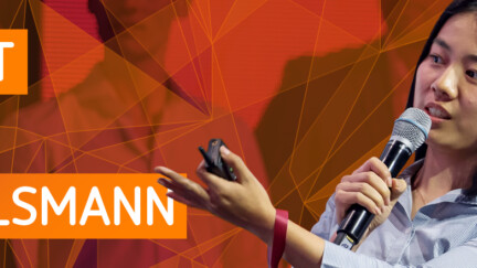 Auf dem Bild sieht man rechts eine Frau mit Mikrofon und ausgestreckter Hand, links vor orange-rotem Hintergrund den Schriftzug Talent Meets Bertelsmann