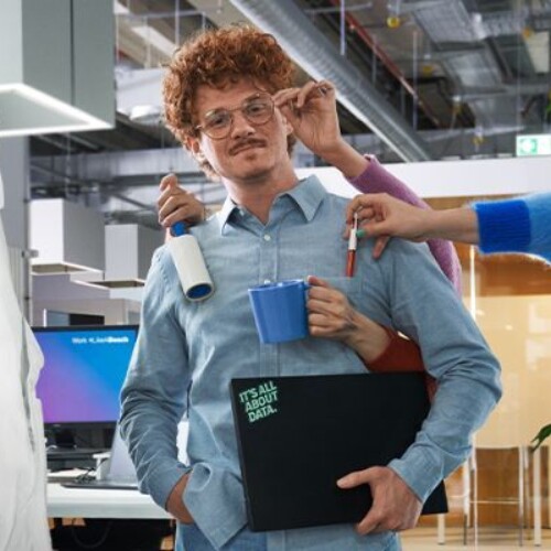 Mann steht mit Laptop in einem Büro. Ihm werden von anderen Mitarbeitern verschiedene Dinge gereicht.