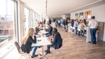 Mitarbeitende sitzen in einem Raum mit großer Fensterfront an Tischen und Essen Brezeln.