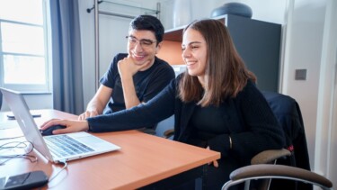 Ein Mitarbeiter und eine Mitarbeiter sitzen gemeinsam am Schreibtisch und schauen auf einen Laptop