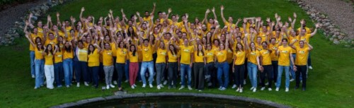 Eine große Gruppe von Mitarbeiter:innen von DHL Consutling streckt die Hände nach oben. Alle tragen gelbeT-Shirts