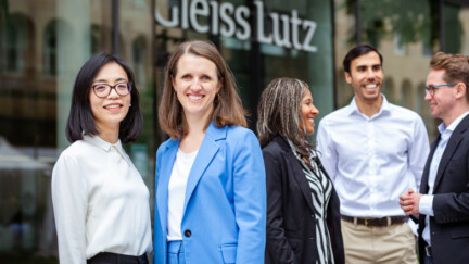 Drei Frauen und zwei Männer im Business-Outfit vor einer Kanzlei mit Glasfassade, auf der Gleiss Lutz steht.