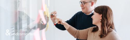 Links unten das Logo von undconsorten, rechts sieht man einen Mann und eine Frau die nach links blicken und auf eine Flipchart schreiben.