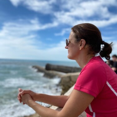 Frau in T-Shirt und mit Sonnenbrille lehnt sich über Mauer und schaut auf das Meer