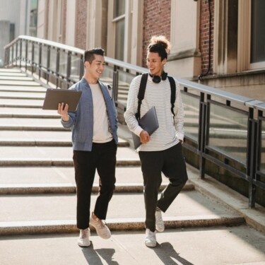 Zwei junge Männer laufen eine Treppe hinunter, dabei unterhalten sie sich. Ein Student trägt eine Mappe, der andere einen Laptop. [Quelle: pexels.com]