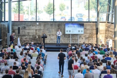 Publikum auf Stühlen sitzend in einer großen Halle, vorne steht ein Redner auf dem Podium