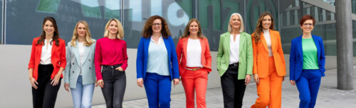 Im Hintergrund des Bildes ist das Allianz-Logo bzw. Schriftzug zu sehen, davor acht Frauen in bunten Hosenanzügen, die nebeneinander stehen.